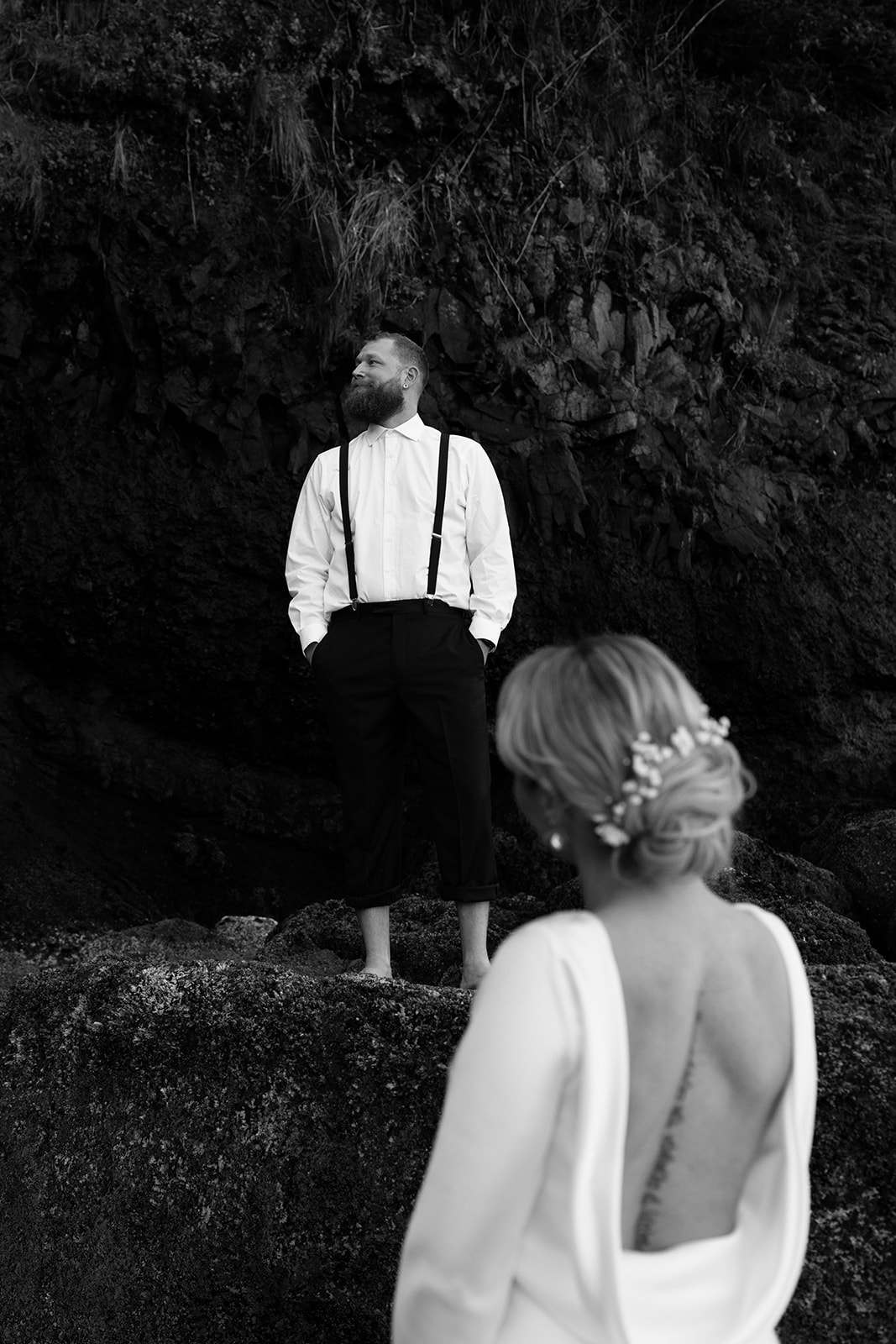 A couple in wedding attire stand near the ocean in front of a rocky black cliff for their Florence, Oregon elopement.