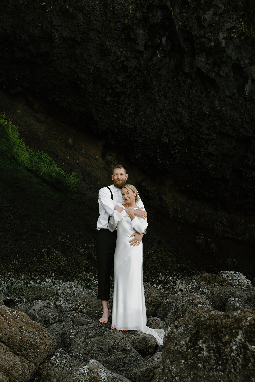 A couple in wedding attire stand near the ocean in front of a rocky black cliff for their Florence, Oregon elopement.