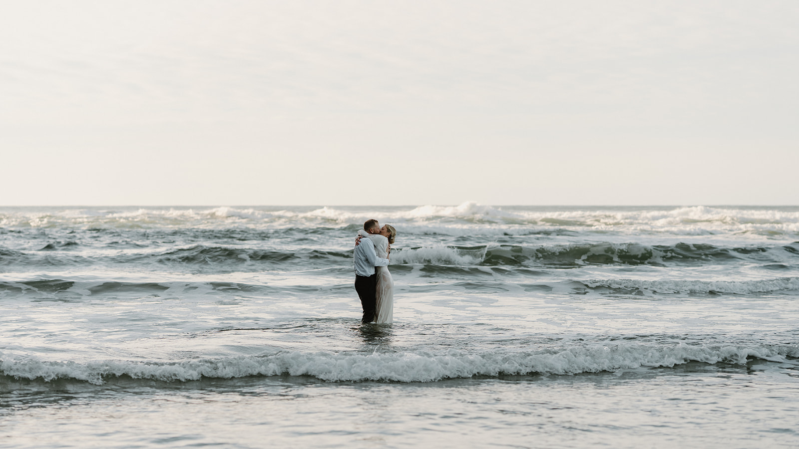 A couple in wedding attire stands small in the ocean with the sun behind them. They embrace and kiss to celebrate their Florence, Oregon elopement. 
