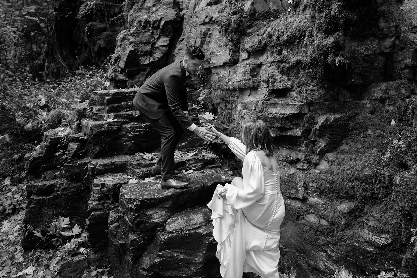 A black and white photo of a groom helping a bride onto a stack of wet rocks during their Latourell Falls elopement.