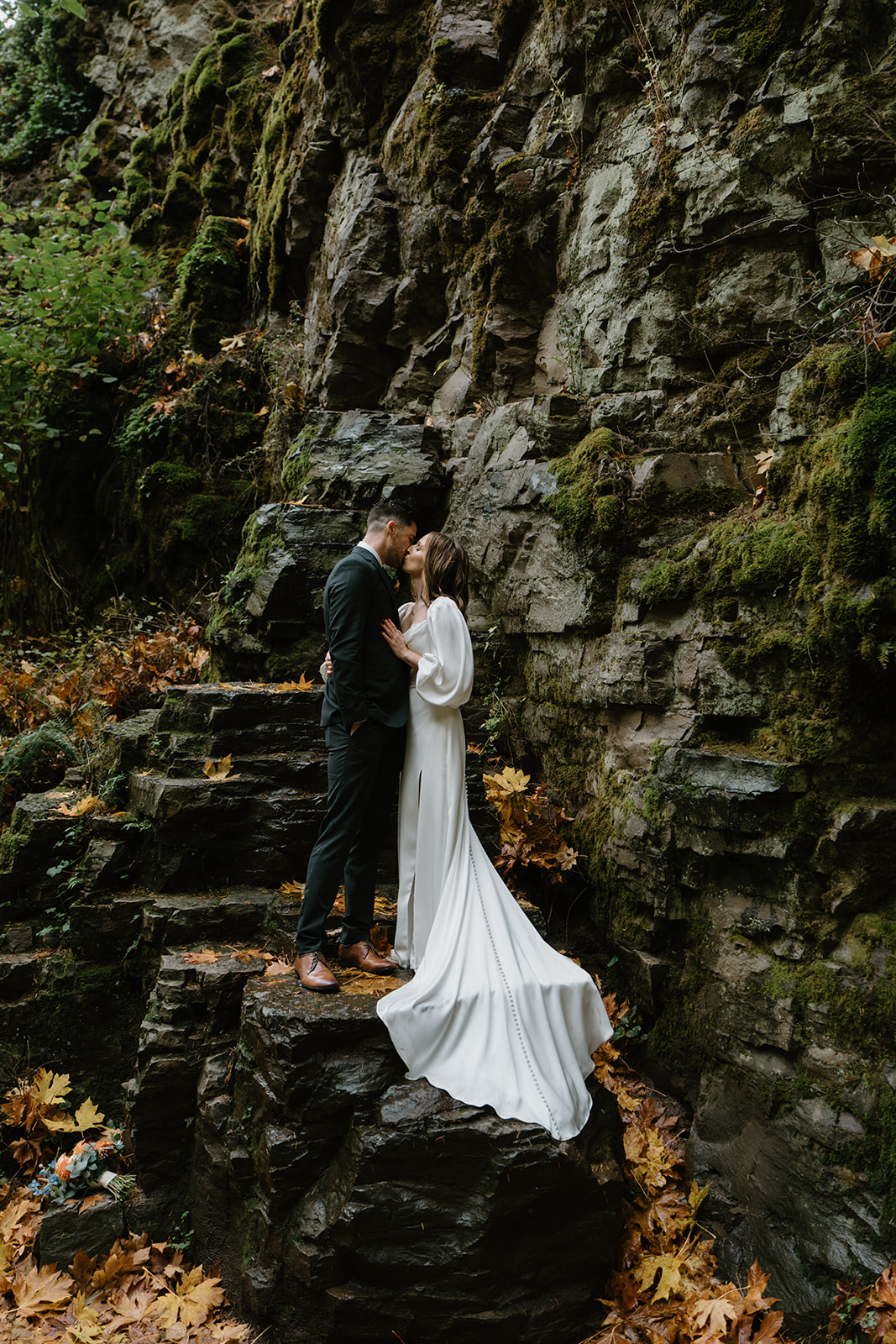 A couple in wedding attire stand on top of wet, black rocks and snuggle in close during their Latourell Falls elopement.