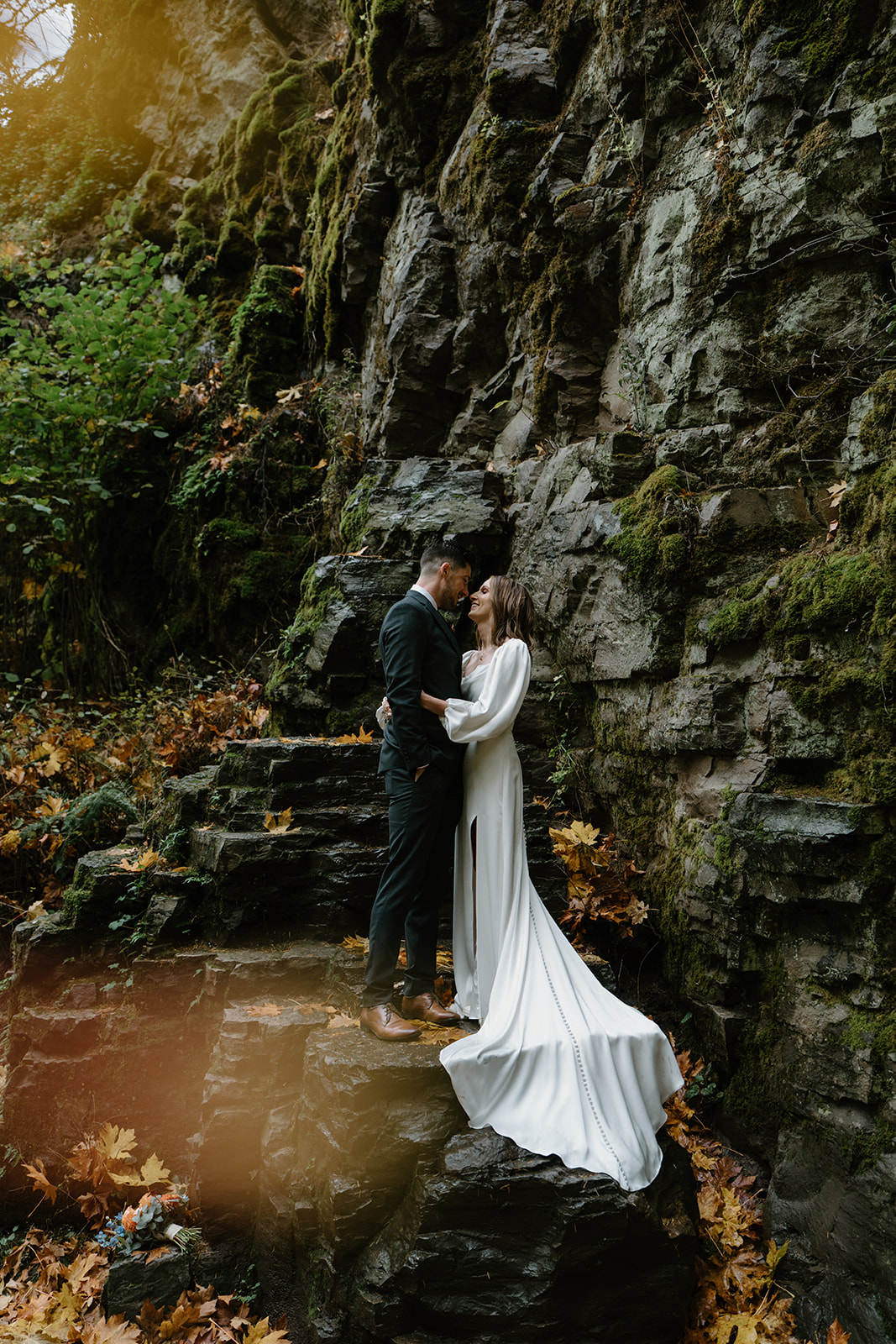 A couple in wedding attire stand on top of wet, black rocks and snuggle in close during their Latourell Falls elopement.