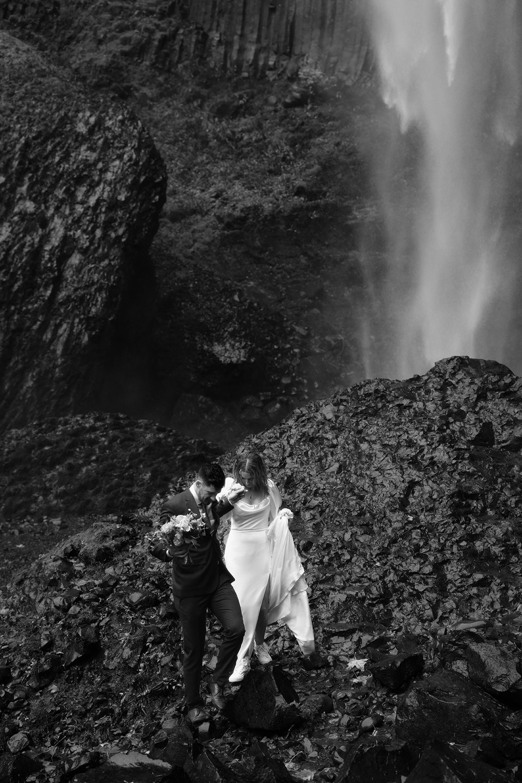 A black and white photo of a couple in wedding attire walking down a wet rock in front of a waterfall during their Latourell Falls elopement.