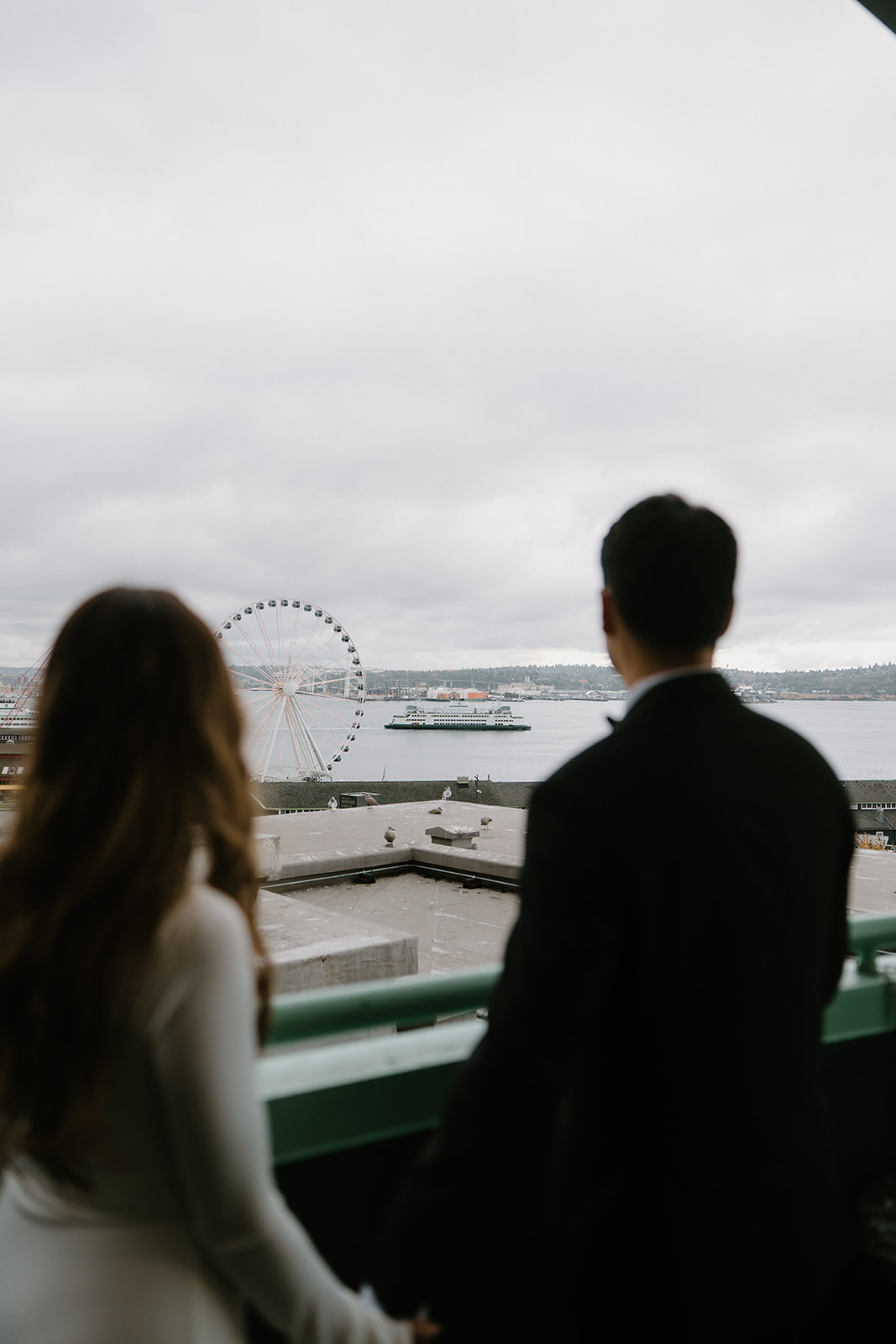 A couple in wedding attire holds hands and looks at a ferris wheel in the distance during their Pike Place elopement. 