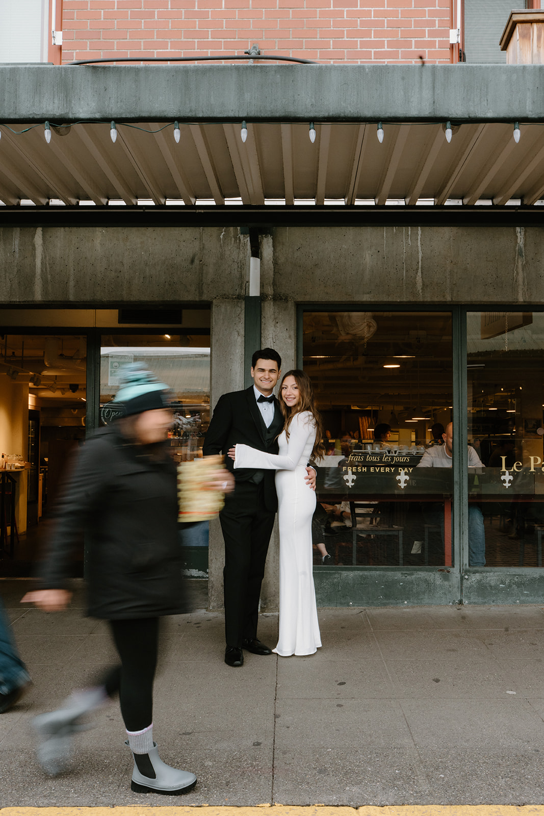 A couple in wedding attire embraces and looks at the camera as people walk by during their Pike Place elopement. 