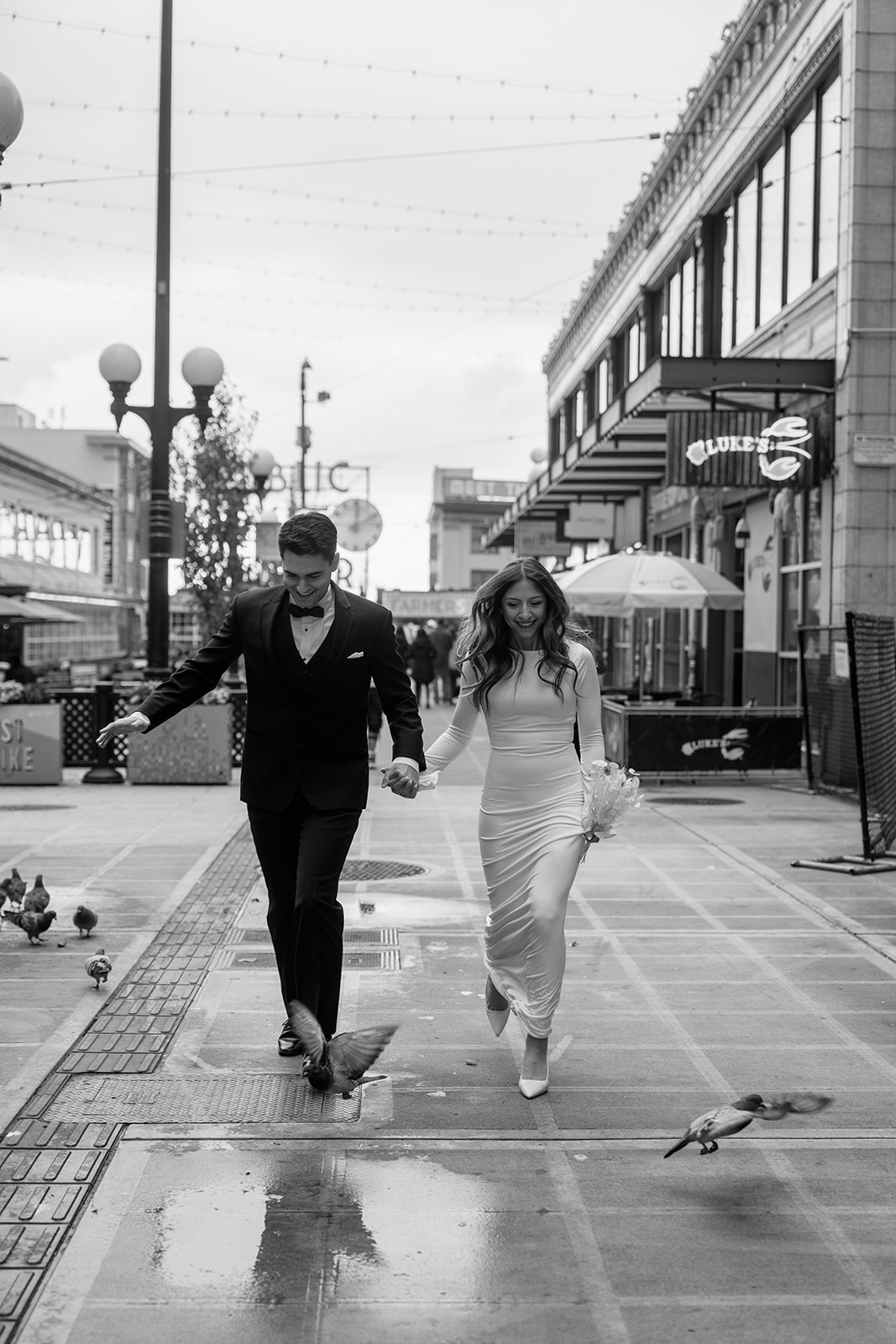 A black and white photo of a couple in wedding attire holding hands and running through a group of pigeons during their Pike Place elopement. 