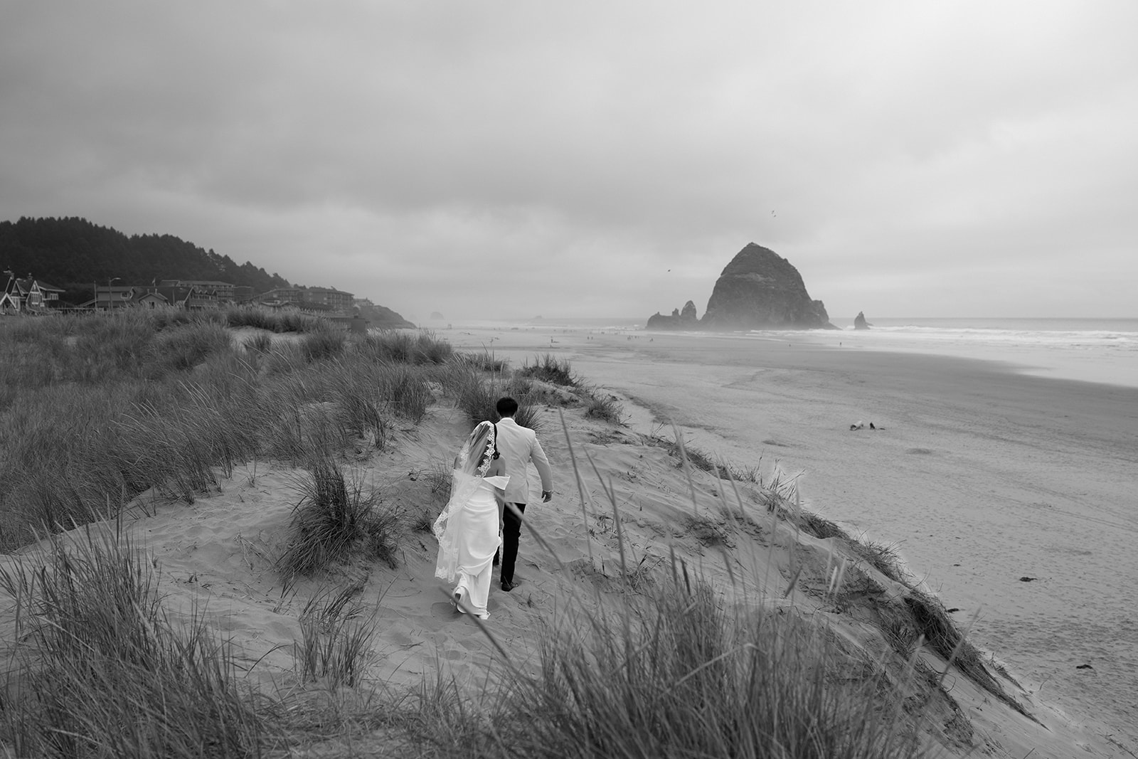A black and white photo of a couple walking in the distance towards Haystack Rock A couple exchanges wedding vows with an officiant in front of Haystack Rock during their Oregon Coast elopement. 