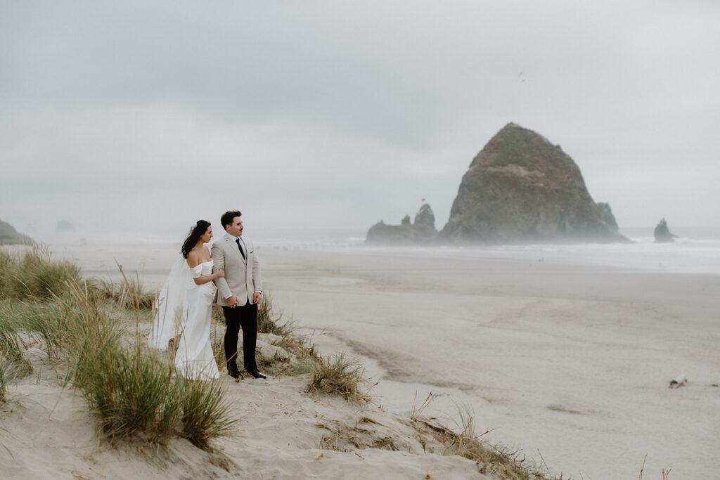 A couple in wedding attire stands on a grassy hill and looks at the ocean A couple exchanges wedding vows with an officiant in front of Haystack Rock during their Oregon Coast elopement. 