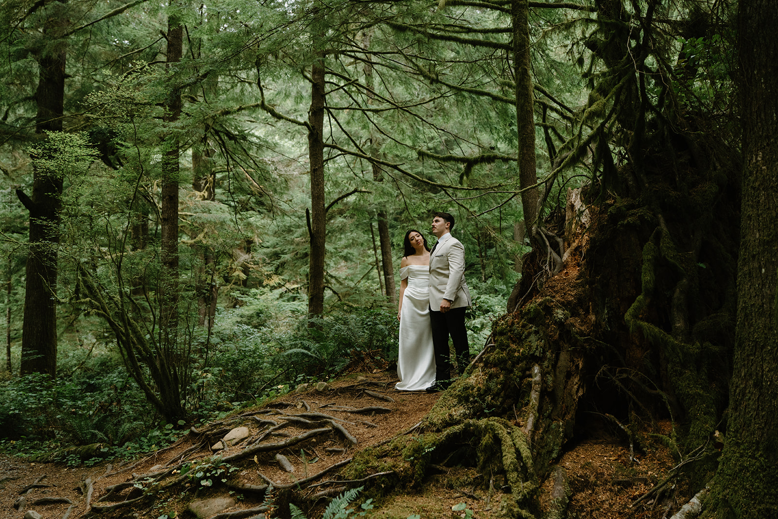 A couple stands in a dark green forest in wedding attire during their Oregon Coast elopement. 