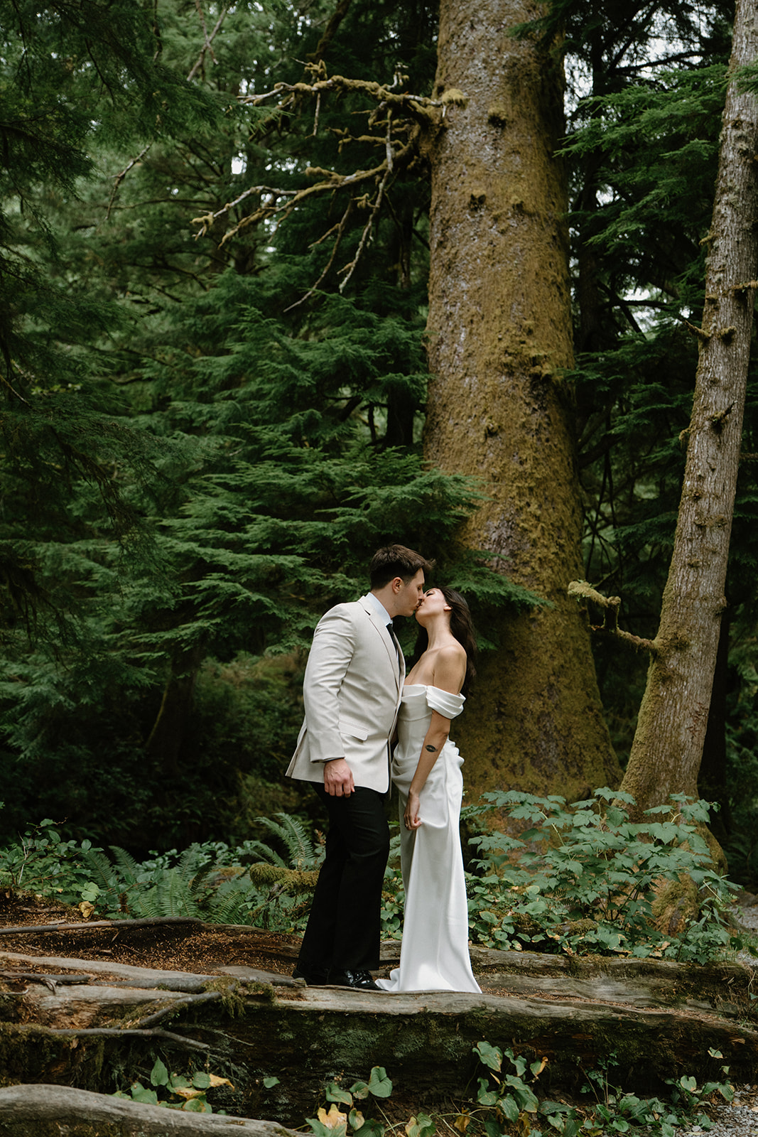 A couple stands in a dark green forest in wedding attire during their Oregon Coast elopement. 