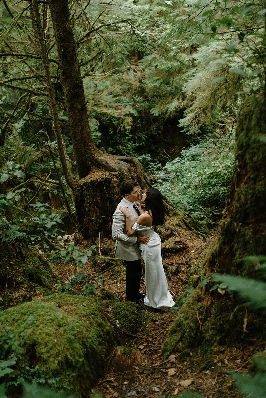 A couple stands in a dark green forest in wedding attire during their Oregon Coast elopement. 