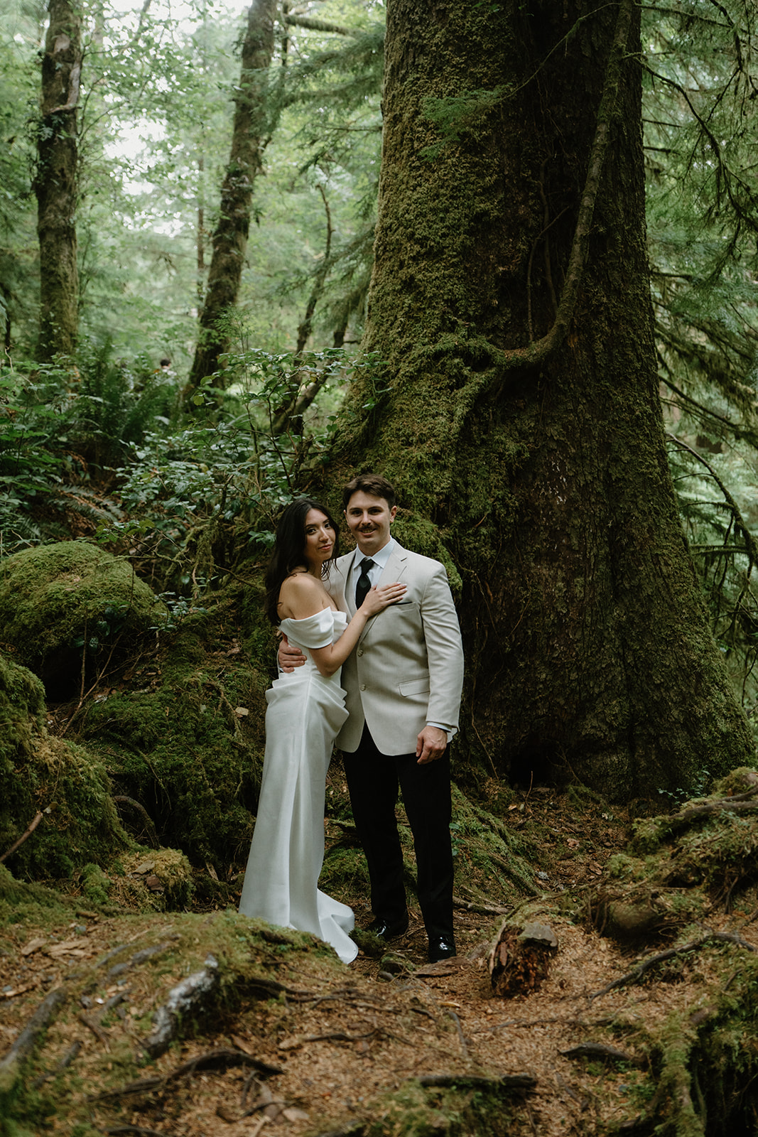 A couple stands in a dark green forest in wedding attire during their Oregon Coast elopement. 