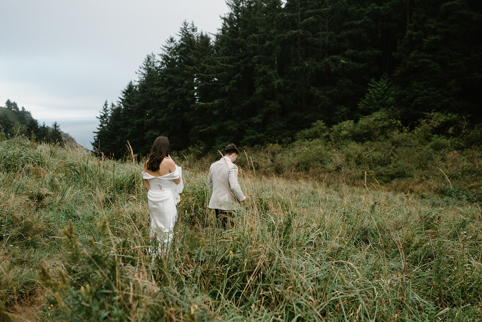 A couple in wedding attire walks through tall green grass during their Oregon Coast elopement. 