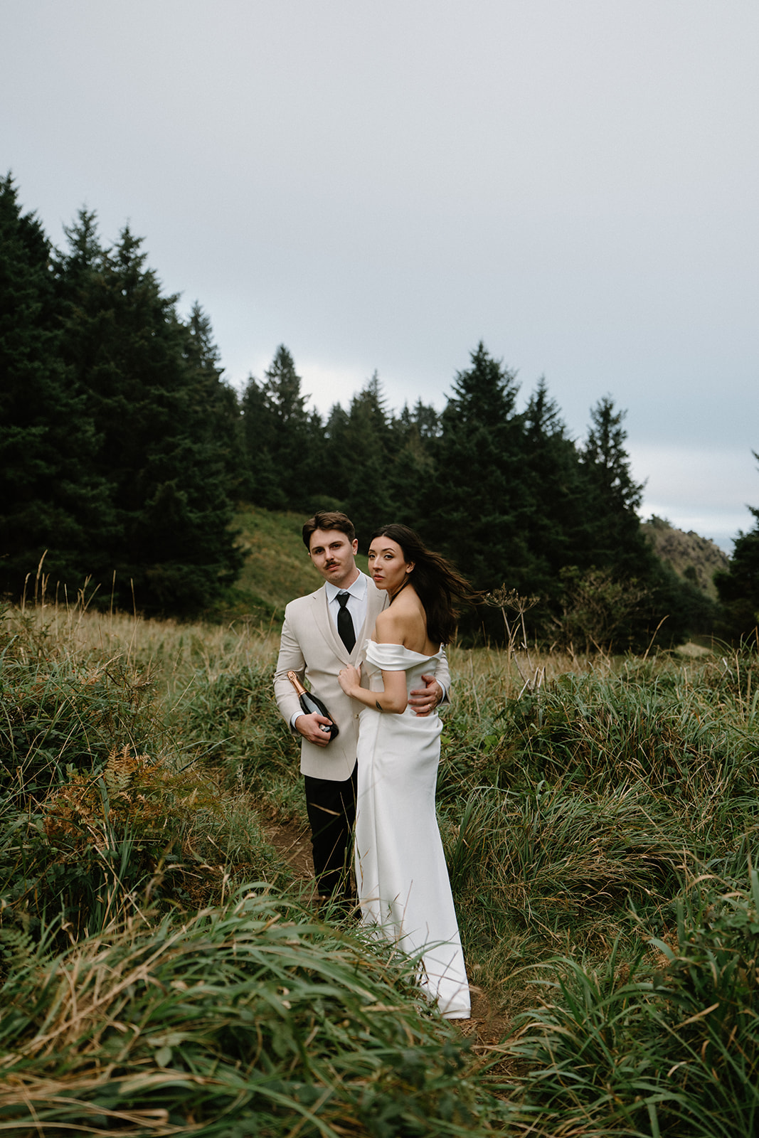 A couple in wedding attire walks through tall green grass during their Oregon Coast elopement. 