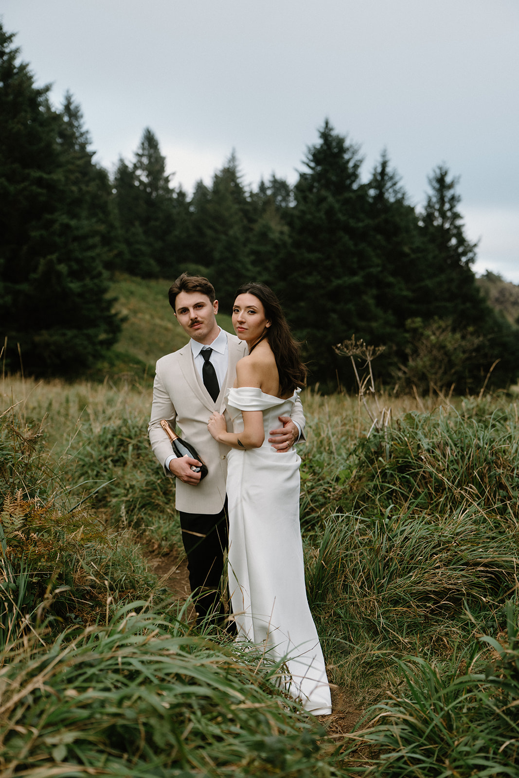 A couple in wedding attire walks through tall green grass during their Oregon Coast elopement. 