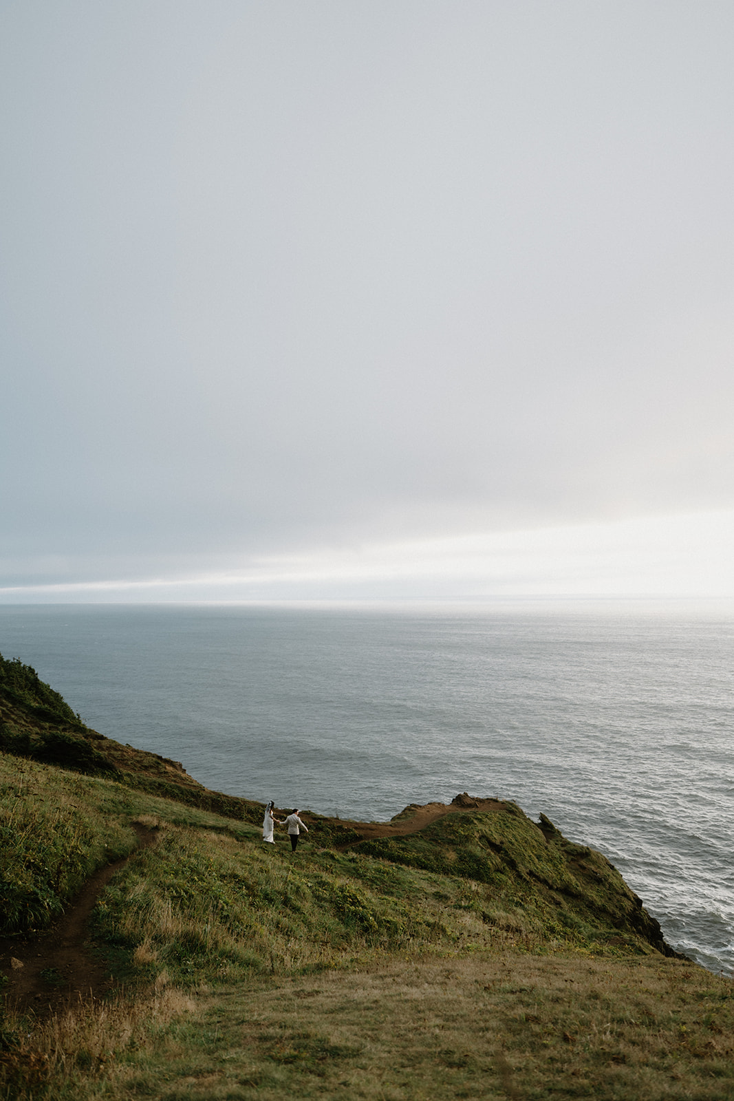 A couple walks far away on the edge of a cliff overlooking the ocean during their Oregon Coast elopement. 