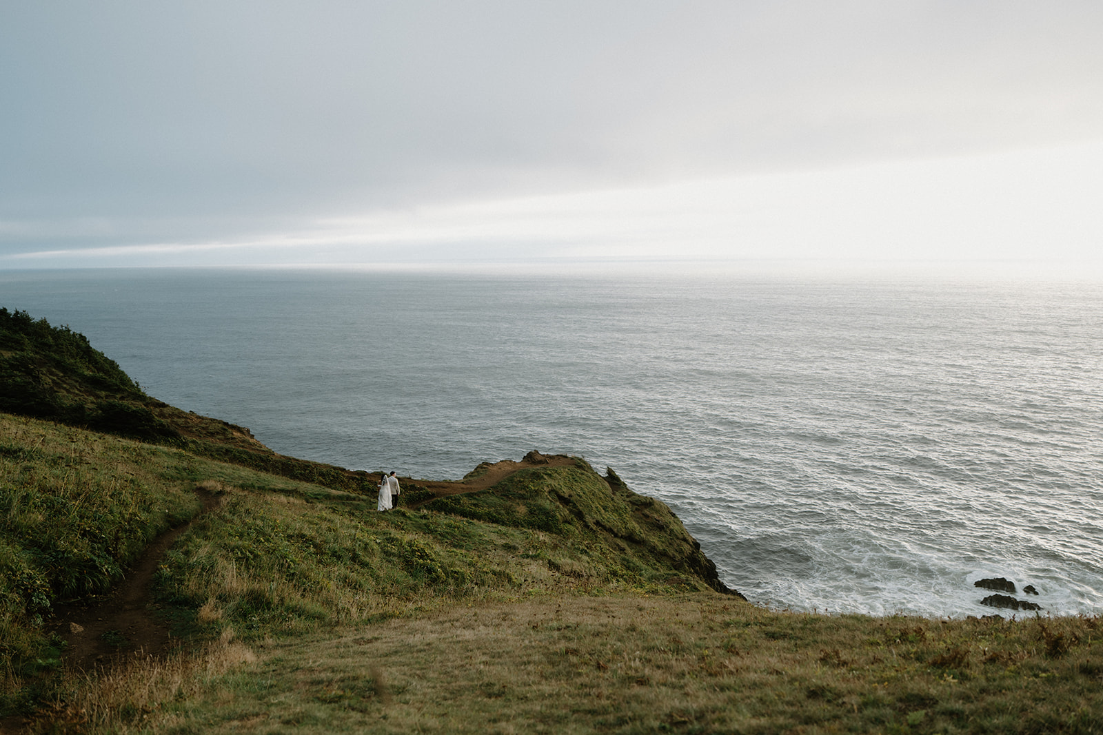 A couple stands extremely small on the side of a cliff overlooking the ocean, during their Oregon Coast elopement. 