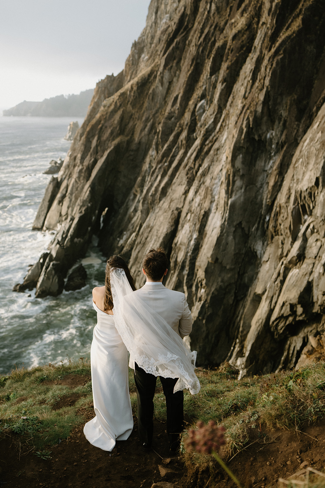 Wind picks up a bride's veil on the edge of a cliff overlooking the ocean during an Oregon Coast elopement. 