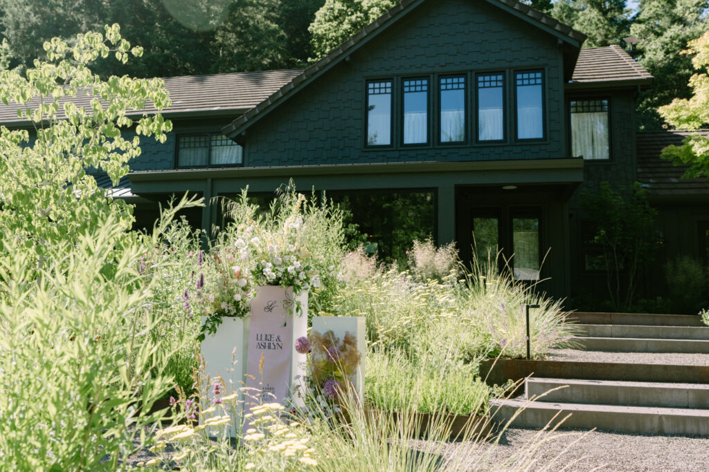 The Carly Estate Wedding venue from the front - a black-ish building sits behind tall grass, flowers, and wedding signage.