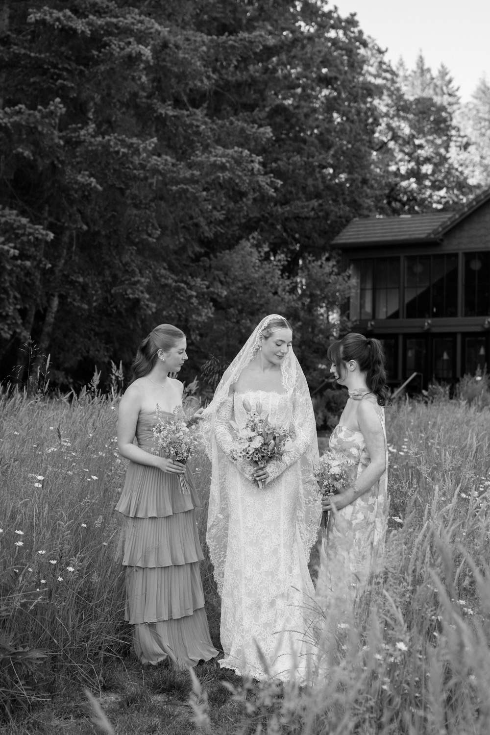 A black and white photo of a bride and two bridesmaids standing in a grass field in front of a modern building during a Carly Estate wedding.