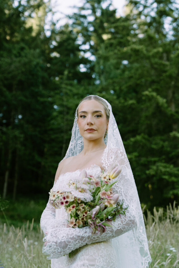 A bride stands and looks down at the camera in a grass field during her Oregon wedding photos.