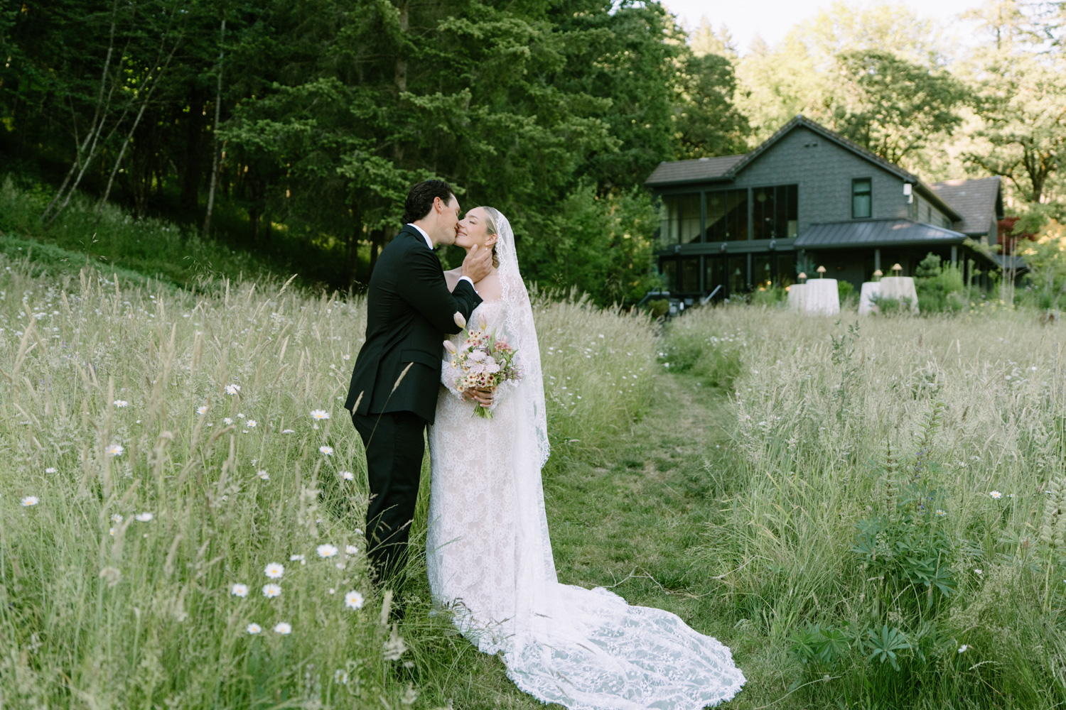 A bride and groom stand close and kiss in a grassy field in front of the Carly Estate wedding venue.