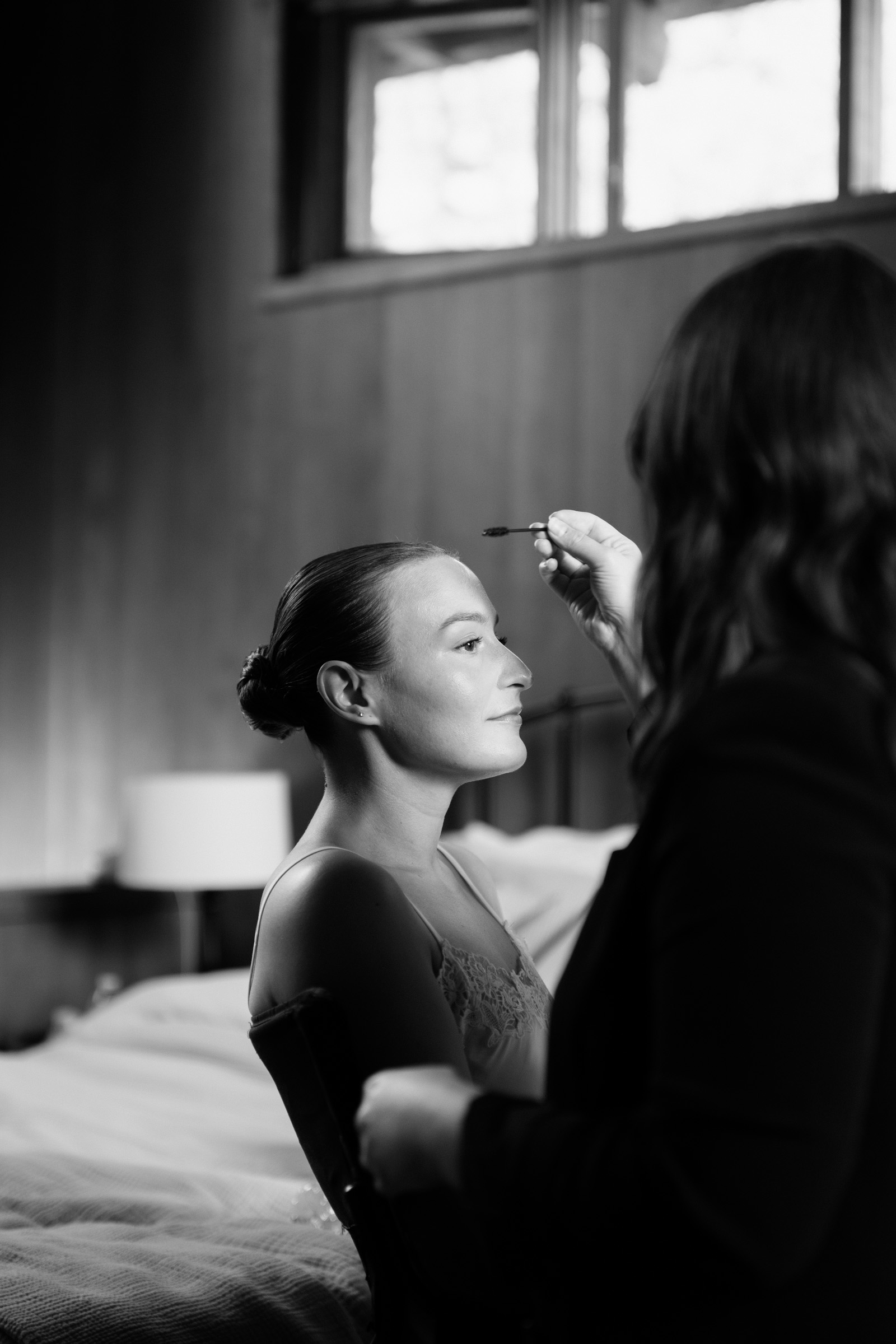 Partner getting wedding makeup done in a wood-paneled room at Anker Place, seated in profile as an artist applies a brush
