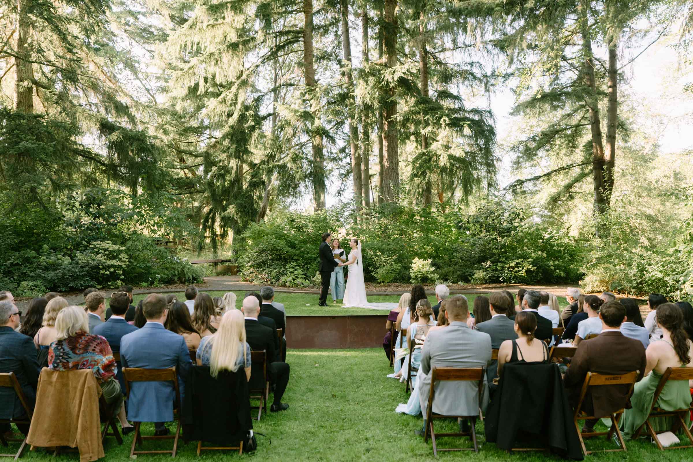 Outdoor wedding ceremony at Anker Place, guests in wooden chairs on a flat lawn clearing ringed by tall evergreen trees