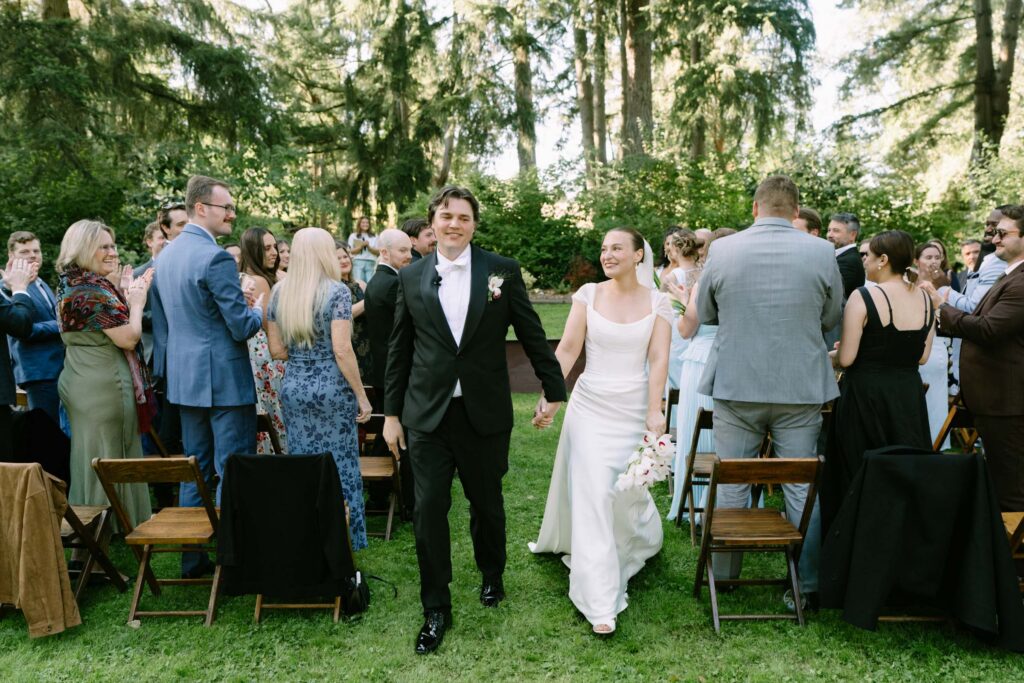 Couple holding hands during the recessional at Anker Place, guests standing among wooden chairs