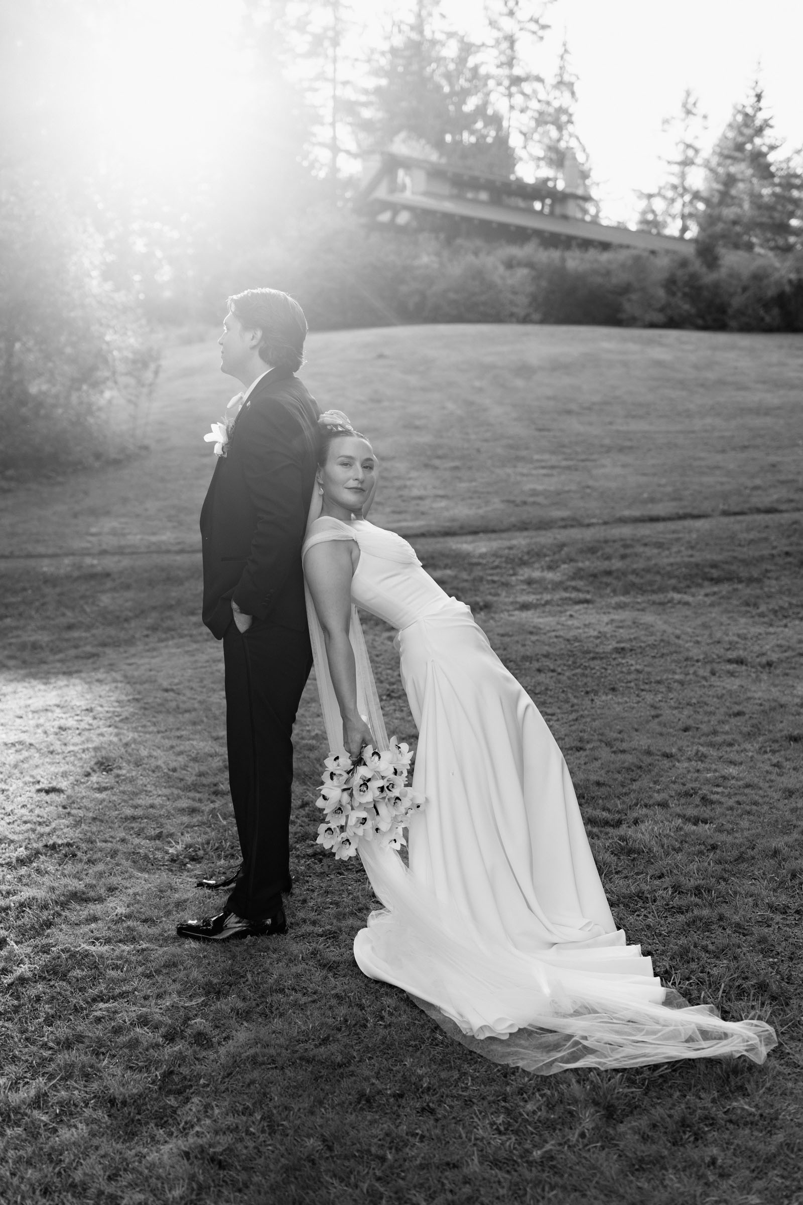 Couple portrait on the lawn at Anker Place, one leaning back with a bouquet against sun flare through the trees