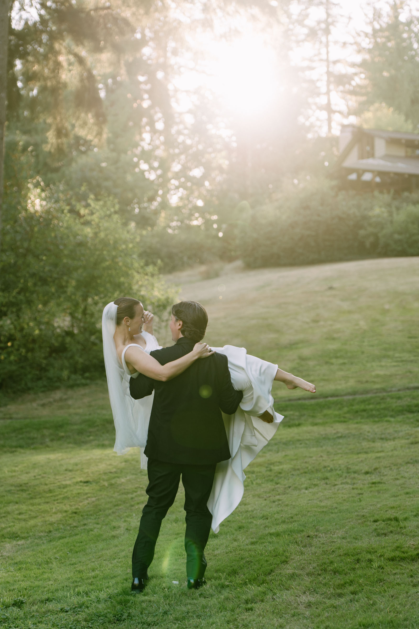 Couple embracing on the lawn near the house at Anker Place, sun flaring through the firs