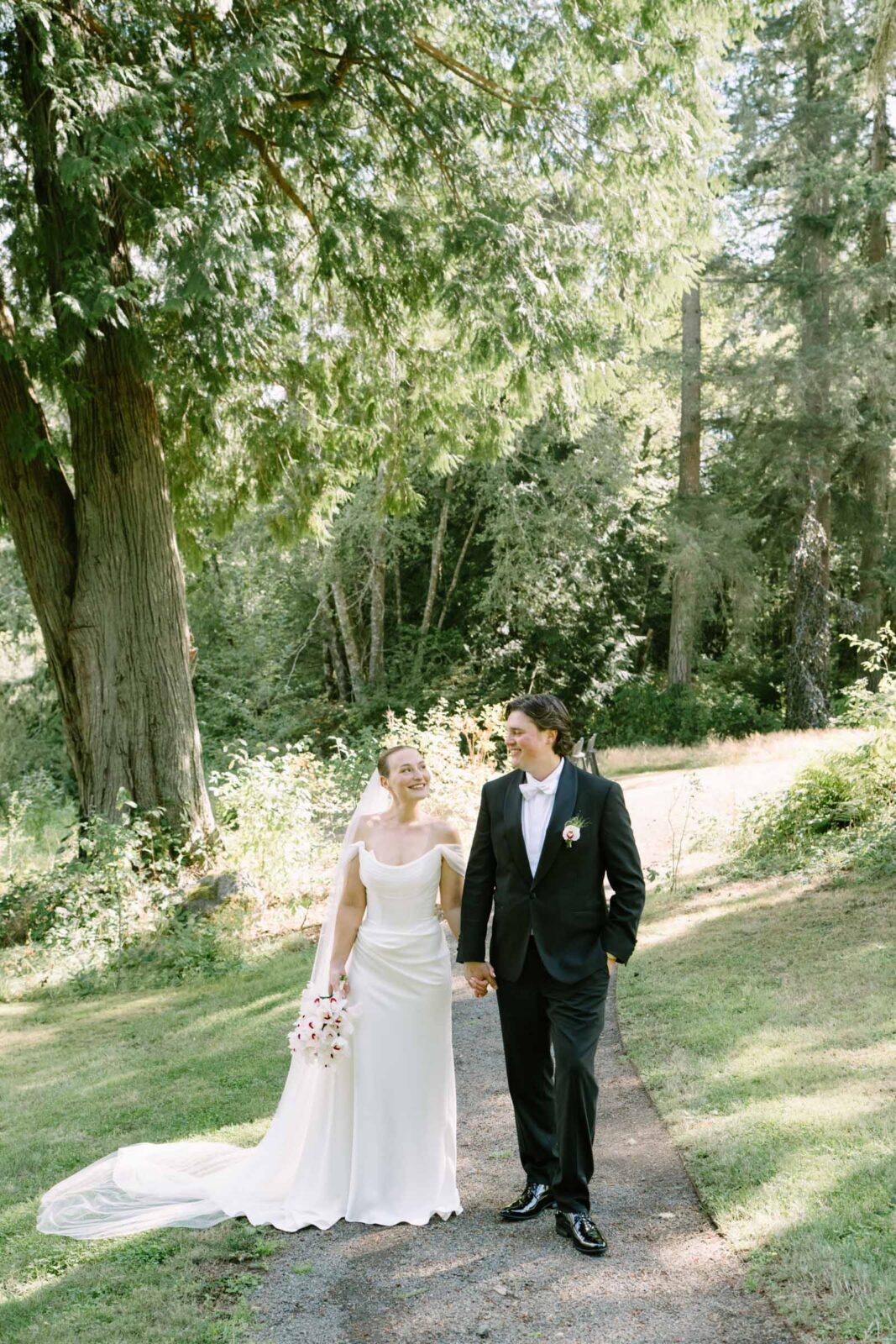 Couple walking a wooded garden path at their Anker Place wedding, gown train trailing behind